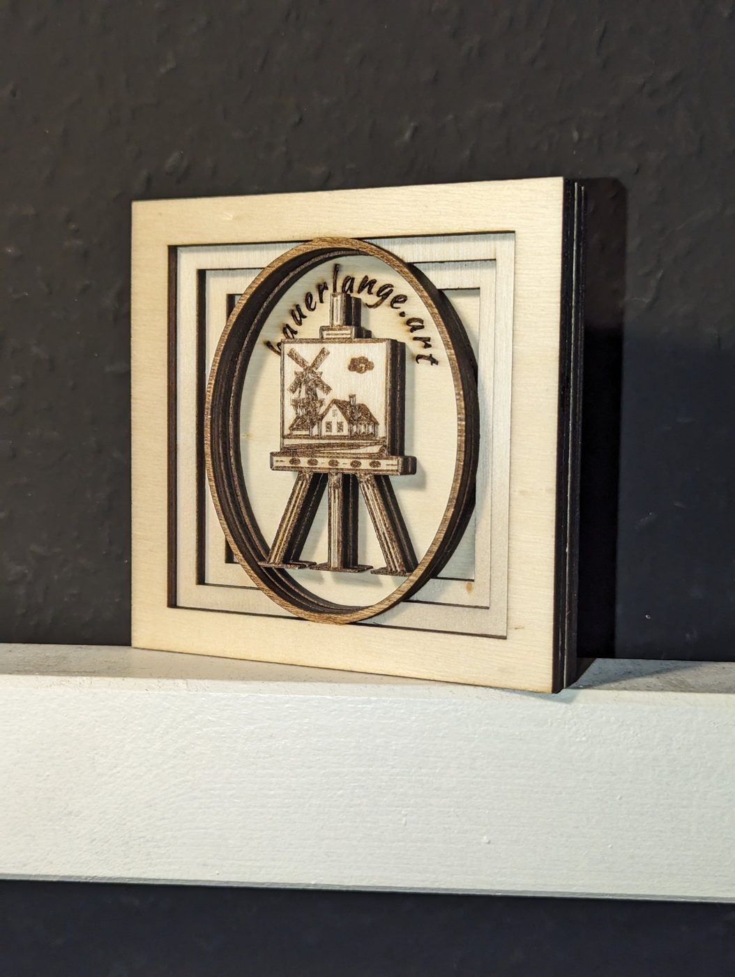 Logo bauerlange.art als geschichtetes Holzrelief: Staffelei mit Windmühle im Oval
